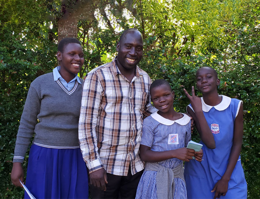 ElijahMillard_s3_1 A group of four people standing outdoors in front of leafy greenery. From left to right, two school girls in uniforms, a man in a plaid shirt, and another school girl in uniform are all smiling; one girl on the right holds up a peace sign, celebrating their recent charity water donation.