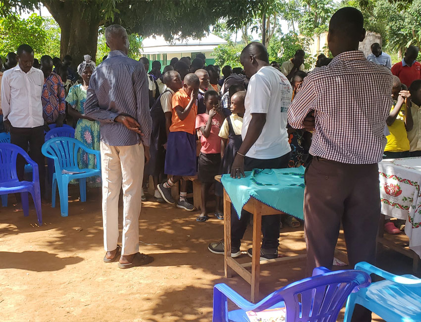 ElijahMillard_s3_3 A group of people, including adults and children, are gathered outdoors under a tree. Several empty blue and purple chairs are in the foreground. A man in a white T-shirt and another in a checked shirt stand near a wooden table covered with cloth, setting up for the charity event with donation boxes nearby.