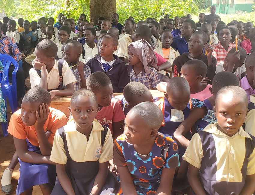 ElijahMillard_s3_4 A group of young students seated closely together on wooden benches outdoors. Many of the children are wearing school uniforms, and some are attentively looking forward while others seem distracted. A tree and greenery can be seen in the background. Nearby, a water station set up through local charity donations supports their daily needs.