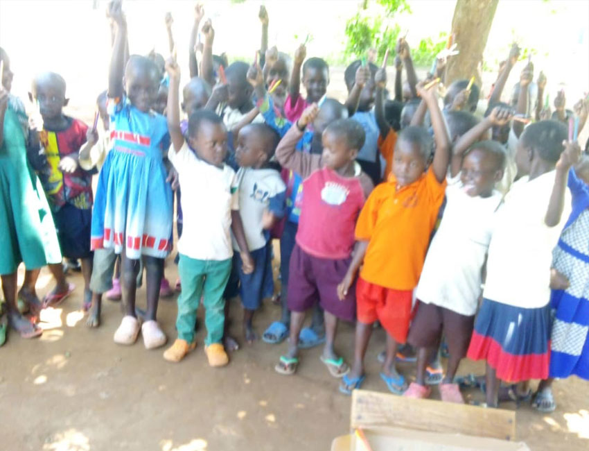 ElijahMillard_s3_6 A group of young children stand outdoors, some with their arms raised enthusiastically. They are dressed in various colorful clothes and appear joyful and engaged. Trees and natural light are visible in the background, suggesting a sunny day. This cheerful scene was made possible through a water charity donation.