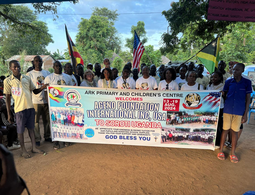2025-Highlights_img1 A group of people, including adults and children, stands outdoors holding a large banner. The banner welcomes the Ageno Foundation International Inc., USA, to Soroti, Uganda. Several flags and text are visible on the banner. Trees and a building are in the background.