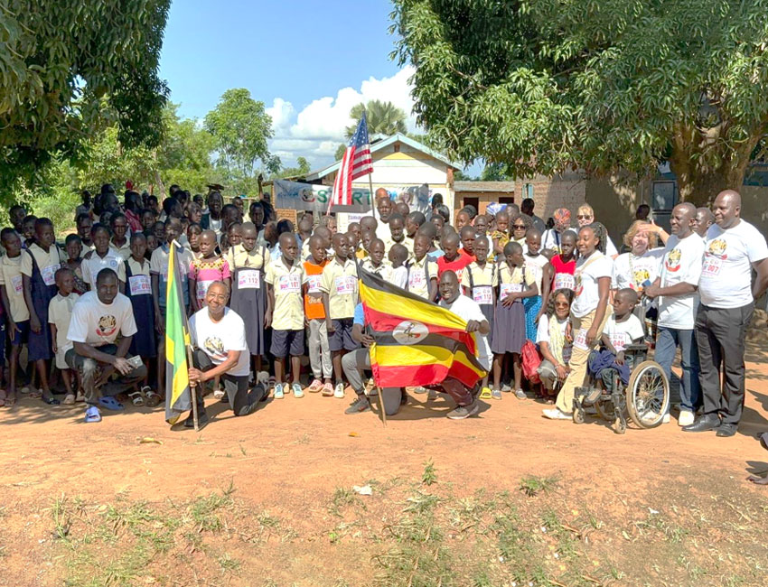 2025-Highlights_img2 A large group of children and adults pose outdoors in front of a building. They hold flags, including Ugandan and American flags. Some wear matching shirts, and one person is in a wheelchair. Trees and a blue sky are in the background.