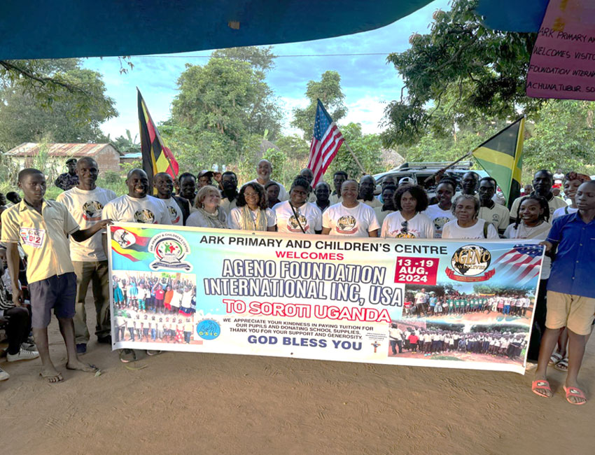 2025-Highlights_img3 A diverse group of people stand outdoors holding a large banner for the "Ageno Foundation International Inc, USA" event in Soroti, Uganda, dated August 13-19, 2024. Flags and trees can be seen in the background.