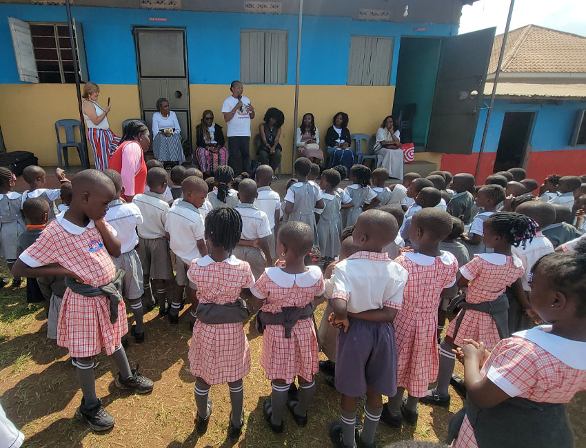 2025-Highlights_img4 A group of young students in uniforms, with checkered dresses for girls and white shirts with gray shorts for boys, are gathered outdoors. They are facing adults, some seated and some standing, addressing them in front of a colorful building.