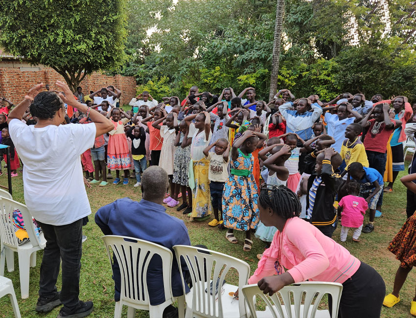 2025-Highlights_img5 A group of children and adults gathered outdoors, following a person leading them in a hand movement exercise. Some people are seated on white plastic chairs. Trees and a brick wall are visible in the background.