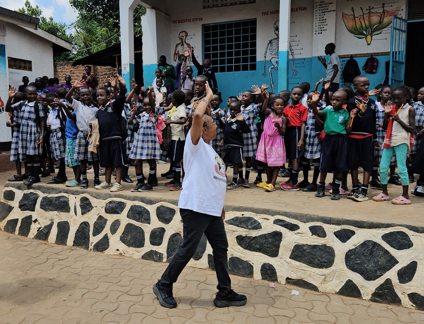 2025-Highlights_img6 A person leads a group of children standing on a platform at a school. The children are wearing uniforms and some casual clothes, waving their hands. The background has educational posters and a mural of a human skeleton.
