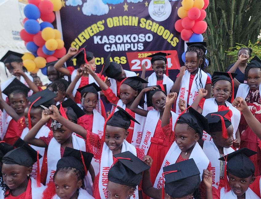 2025-Highlights_img7 A group of children wearing graduation caps and sashes celebrate at a graduation event. They stand in front of a colorful balloon arch. A banner reads "Clever's Origin Schools, Kasokoso Campus, 1st Class 2024.