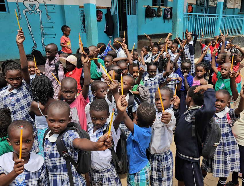 2025-Highlights_img8 A group of schoolchildren in plaid uniforms hold up pencils enthusiastically. They are gathered outside, standing closely together, with a blue building in the background.