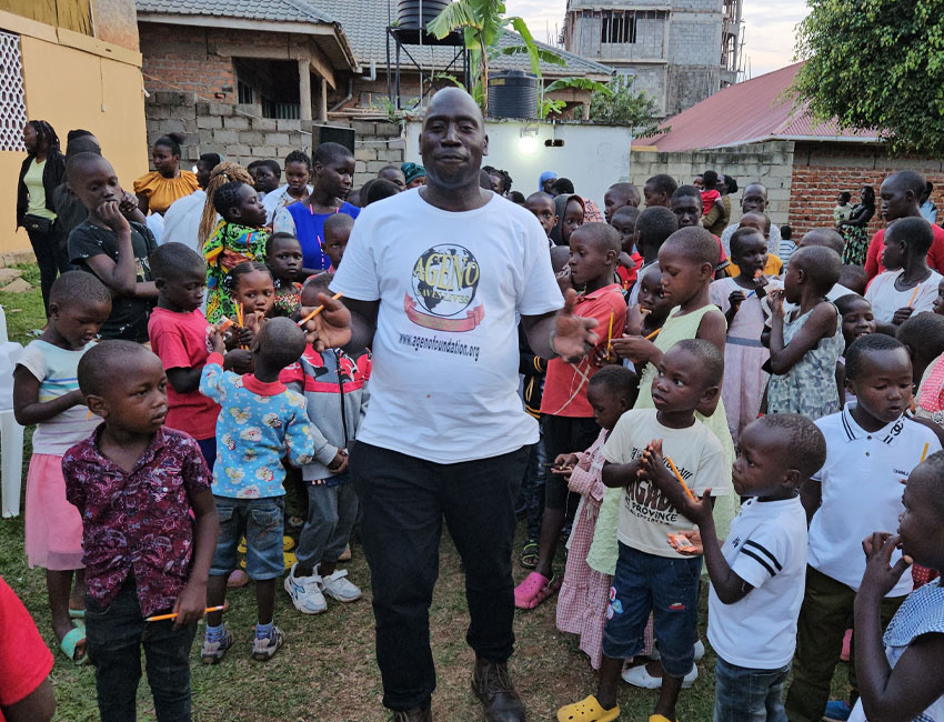 2025-Highlights_img9 A man in a white t-shirt stands smiling among a group of children outdoors. The children, of various ages, gather around him. Some people are standing in the background near buildings and vegetation.