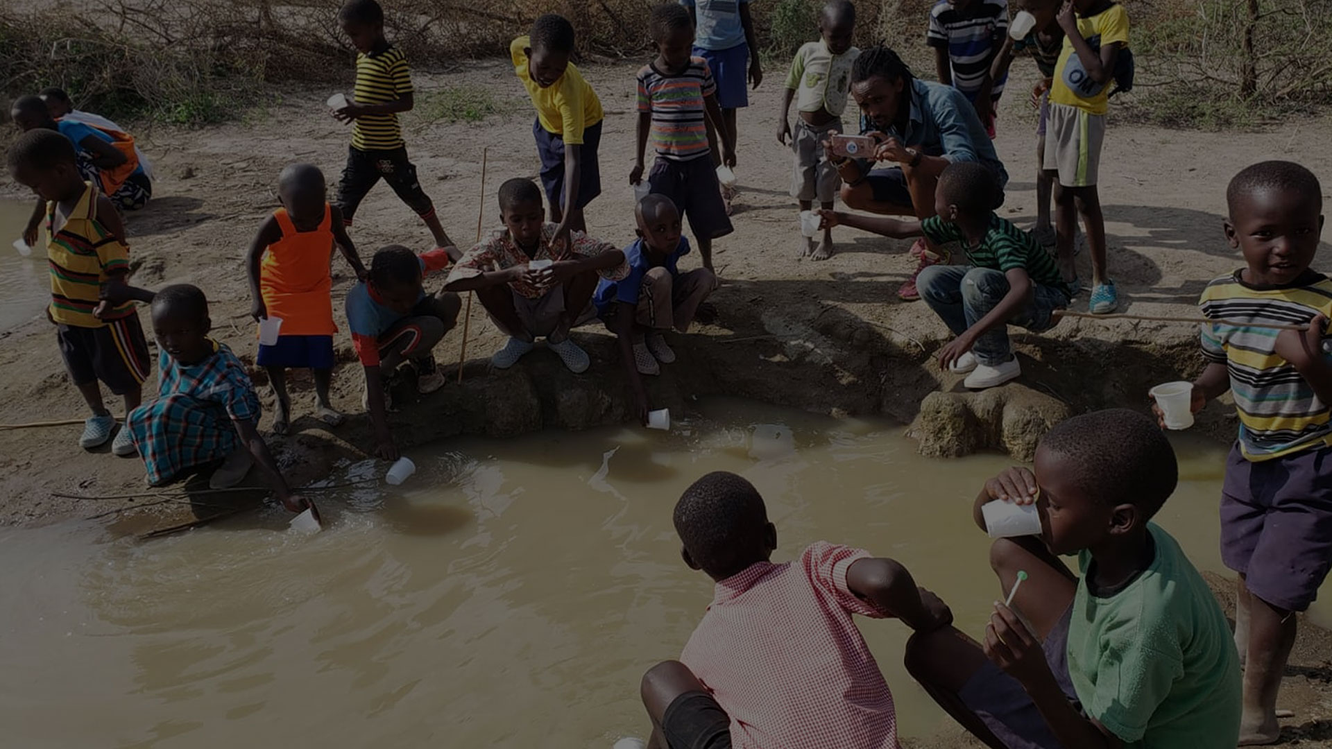 A group of children gather around a muddy waterhole. Some are filling containers with water while others drink from cups. An adult, possibly a veterinarian, is among them, aiding the children. The background shows a dry, rural landscape with sparse vegetation.