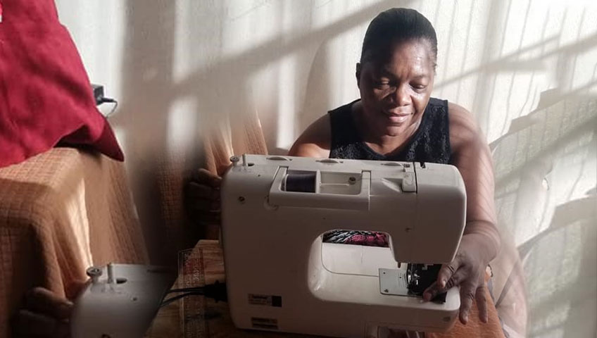 A woman sits at a sewing machine in a softly lit room, intently working on a piece of fabric she plans to donate. She is wearing a dark sleeveless top and appears focused on her task, with sunlight casting gentle shadows around her.