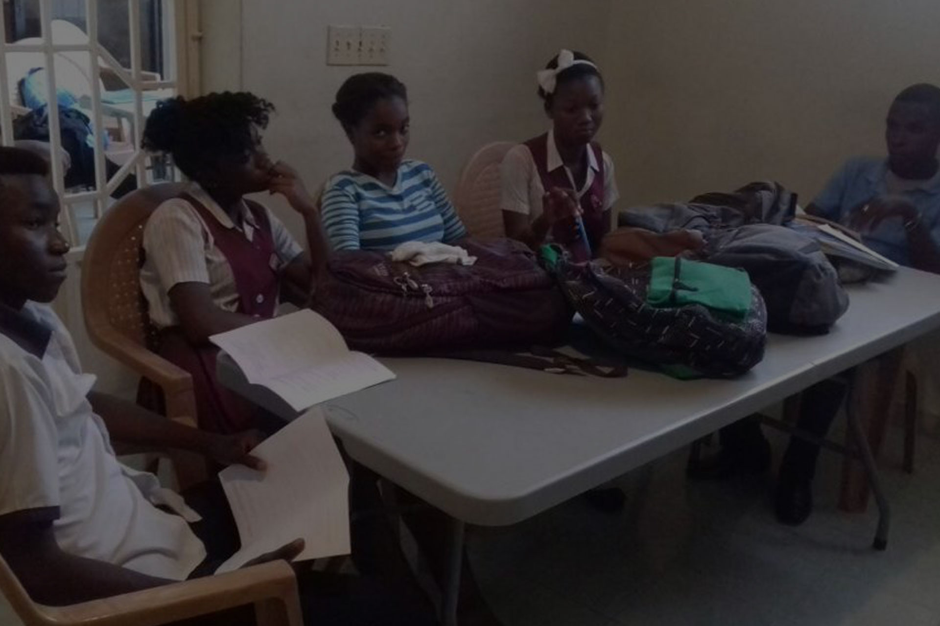 A group of five students, dressed in school uniforms, sit around a rectangular table in a classroom. They are engaged in discussion about organizing a charity event, with books, papers, and backpacks spread out on the table. The room is dimly lit, and there are chairs and a door in the background.