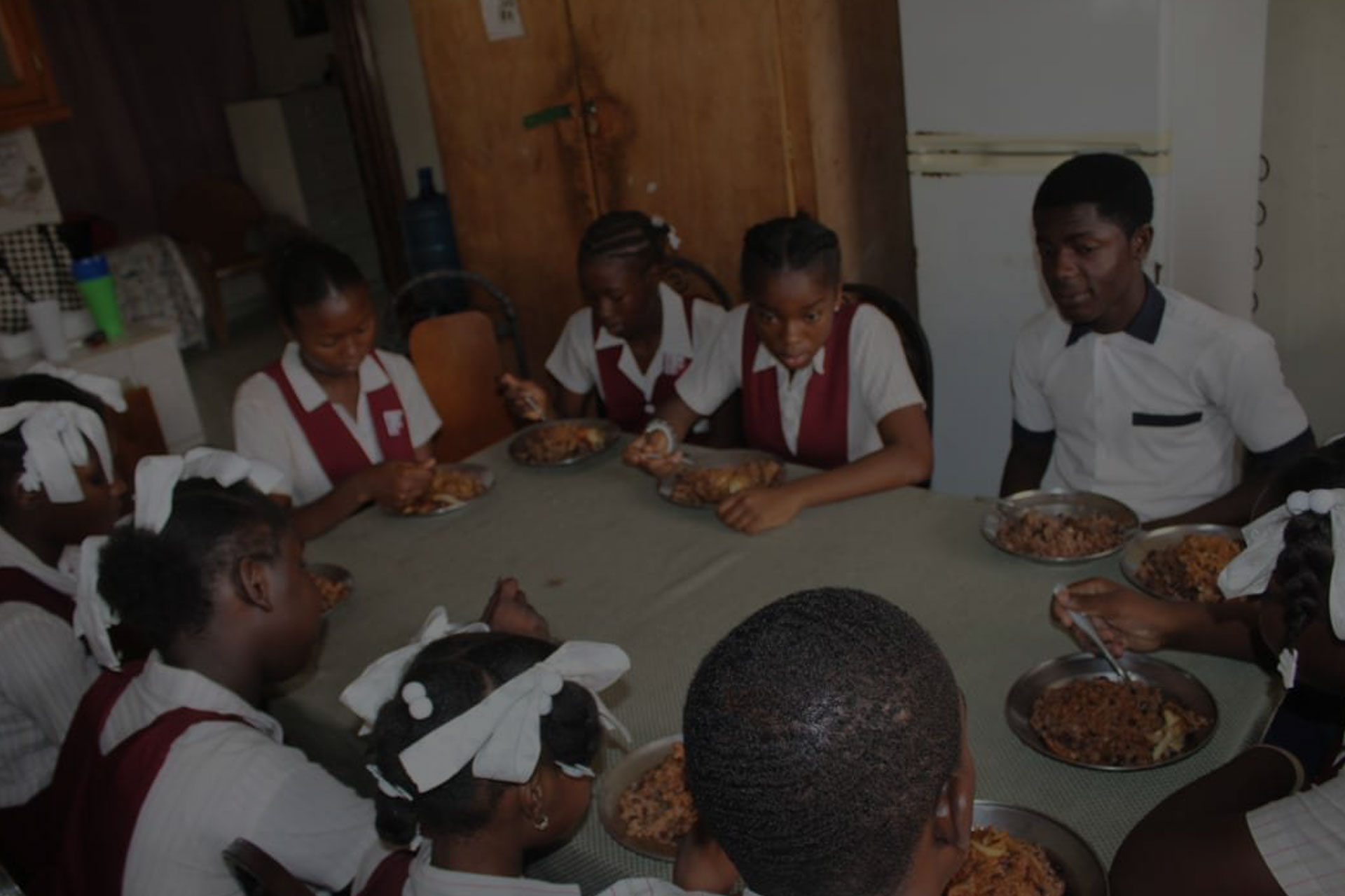 A group of school children in uniforms and a teacher are seated around a table, eating a meal. The children are wearing white shirts, red vests, and white hair ribbons. A jug of water is on the table. The setting appears to be a dining area with wooden cabinets and a refrigerator in the background.