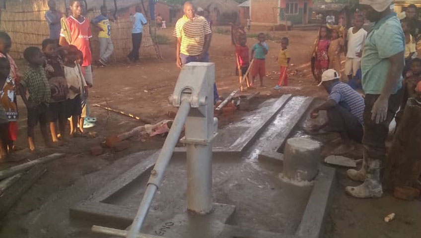 A group of people, including children, gather around a newly constructed concrete water pump in a village setting. Some individuals are working on completing the installation, while others observe. The project was made possible through generous donations. Huts and other buildings are visible in the background.