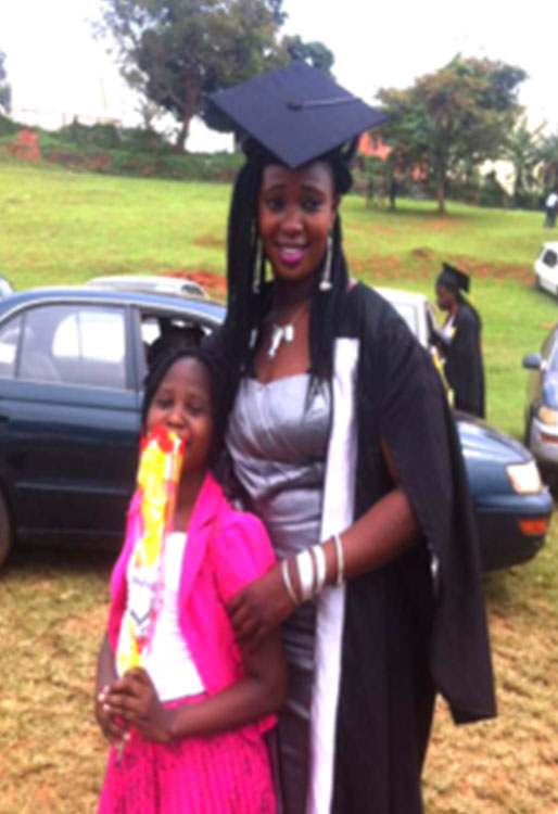 A person wearing a graduation cap and gown stands outside, smiling, with an arm around a child who is holding a colorful toy. There are several cars and trees in the background, celebrating their achievement as a newly minted veterinarian.