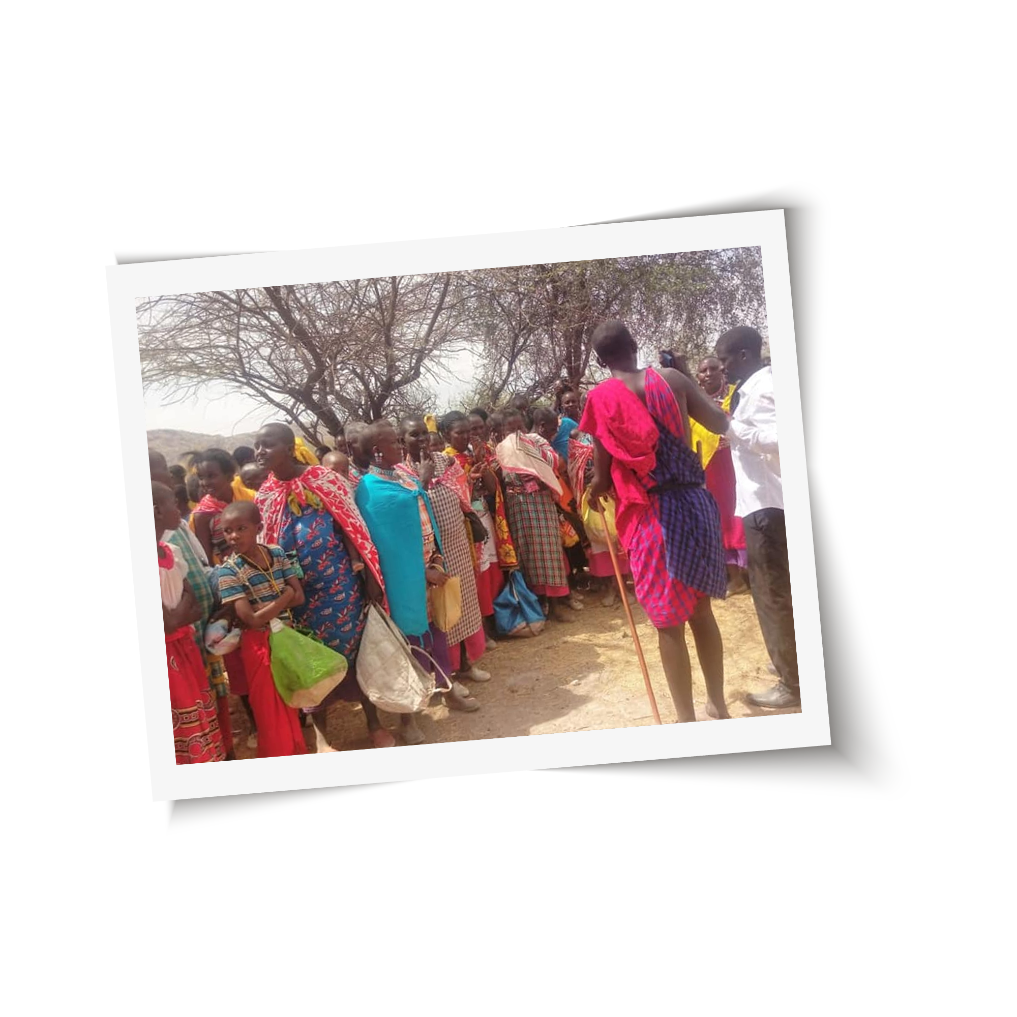 A group of people dressed in colorful traditional garments gather outdoors, with a few individuals holding bags. They are standing beside leafless trees under a clear sky, and their attire and scenery suggest a community event in a rural area, perhaps attended by the local vet as part of the celebration.