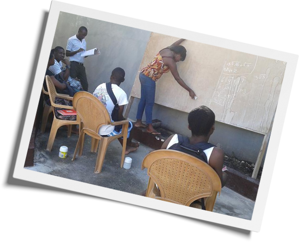 A group of people are gathered outdoors for a class. One person, possibly a veterinarian, stands at a chalkboard, writing, while four others are seated on plastic chairs, attentively watching and taking notes. A man in the background holds a notebook. The setting appears informal.