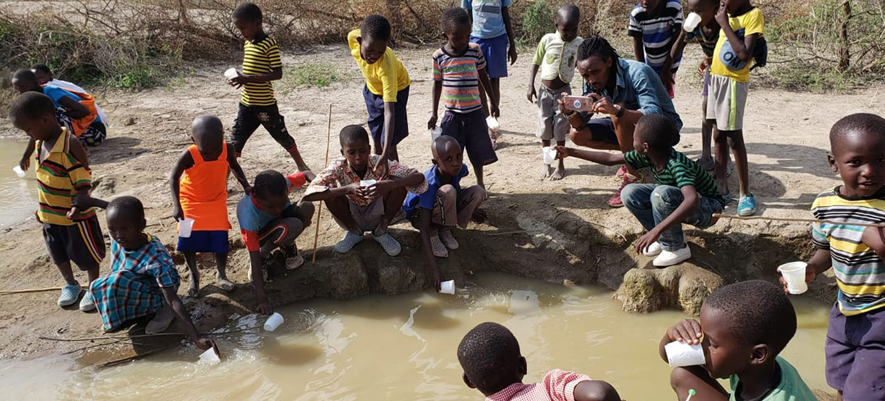 A group of children and an adult gather around a small pond of murky water. Some children scoop water with cups, while others look on. The adult, a vet, appears to be testing the water using equipment. Trees and dry land are visible in the background.