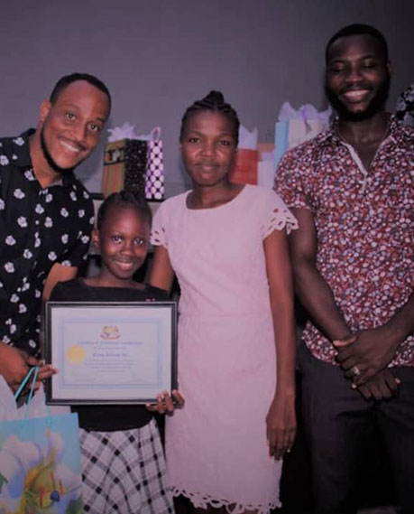 A group of four people stands together smiling. One person, a child, holds a framed certificate—recognition for their donation efforts. Two people on the left are men, and the two on the right are women; one woman is a child. Gift bags and wrapped presents are visible in the background.