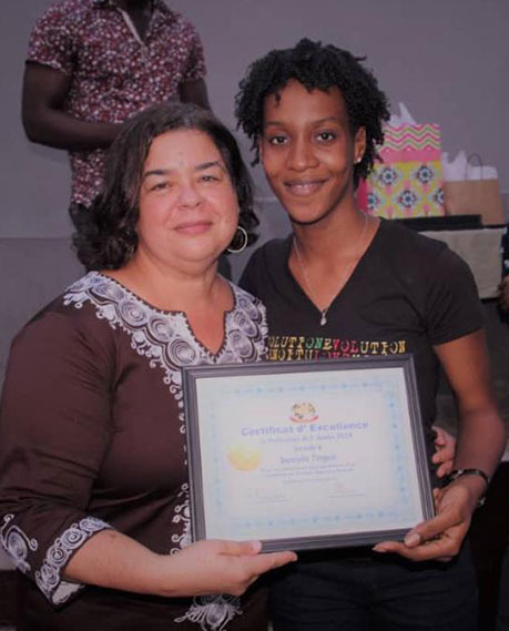 Two people are posing for a photo. One person is holding a framed Certificate of Excellence. The individual on the left, involved in charity work, is wearing a brown outfit with white embroidery, while the person on the right is in a black T-shirt. They are smiling and standing close together.