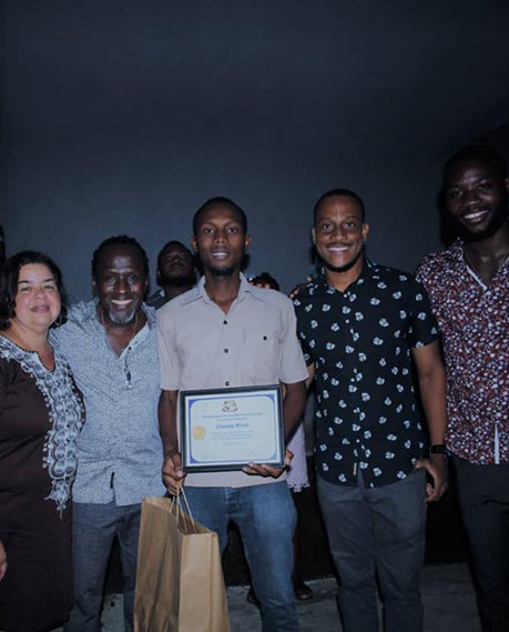 A group of six people stands closely together, smiling at the camera. The person in the middle holds a certificate and a brown paper bag, indicating their donation to charity. The background is dark, which makes the group and their expressions the focus of the image.