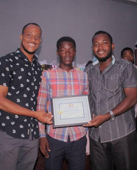 Three men are posing for a photo indoors. The man in the center is holding a framed certificate. The man on the left, who made a generous donation, is wearing a black patterned shirt, and the man on the right is wearing a gray shirt. They all look happy and relaxed.