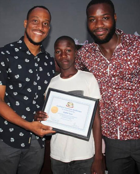 Three men are posing together for a photo. Two adults are standing on either side of a teenager, who is holding a framed certificate from a recent charity event. All three are smiling, and they appear to be indoors in front of a dark background.