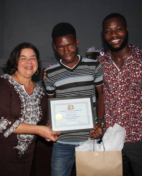 A woman and two men pose for a photo. The woman on the left, wearing a brown and white dress, holds a certificate with one man in the center, who accepts it. The second man, on the right, wears a patterned shirt and holds a gift bag. All are smiling after making a donation to charity.