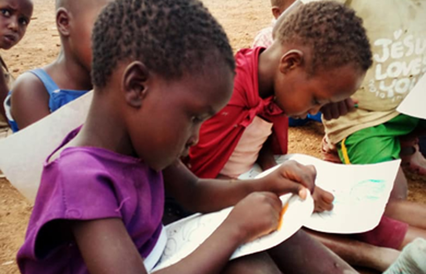 Several young children sit closely together on the ground, focusing intently on drawing or coloring on sheets of paper. They are outdoors near a sandy area. The scene conveys a sense of concentration and creativity, as if inspired by a charity event where donations support their access to art supplies and clean water.