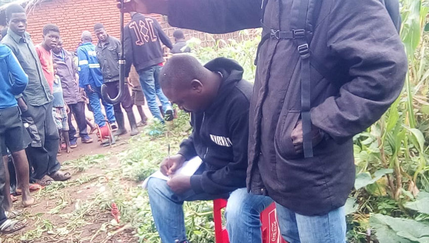 A man sits on a red plastic chair writing on a paper, wearing a hooded sweatshirt. Another person stands nearby, partially in view. Several people stand in the background on a dirt path near a donation station, with a brick wall and plants on either side of the path.