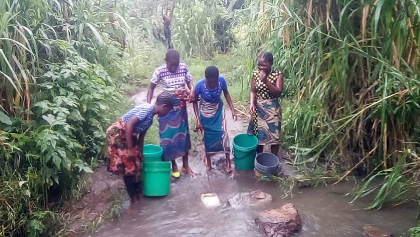 Four women are collecting water from a small stream in a lush, green area. They are wearing colorful dresses and are using plastic buckets and containers to gather the water. Tall grass and vegetation surround the stream, emphasizing the crucial need for clean water donations to aid communities like these.