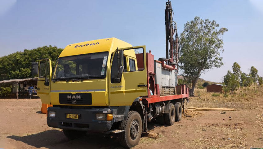 A yellow and red drilling truck labeled "Everfresh" is parked on a dirt road, equipped with a tall drilling rig for water extraction. There are trees and a small hut in the background under a clear blue sky.
