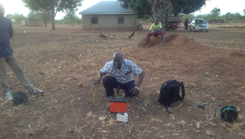 A man in a checkered shirt kneels on the ground, working with electronic equipment outdoors. There is dry, brown grass and a tree behind him, highlighting the need for water. Another man sits by the tree. A house and a parked car are visible in the background.