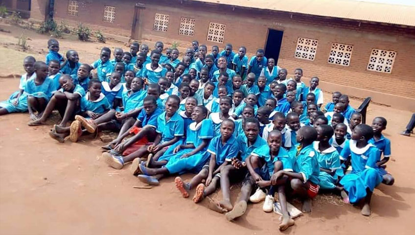 A large group of school children, dressed in blue uniforms, are sitting closely together on the dirt ground outside a brick building. The children, mostly smiling, appear to be of elementary school age. The building in the background has several windows with white latticework, made possible by a generous charity donation.
