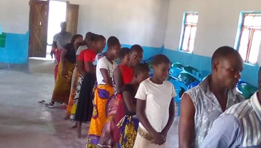 A group of people, including men and women in bright clothing, are standing in a single-file line inside a spacious room with blue and white walls. Nearby, blue plastic chairs are arranged in the background, and an open door is visible. They appear to be waiting to donate water supplies for a community drive.