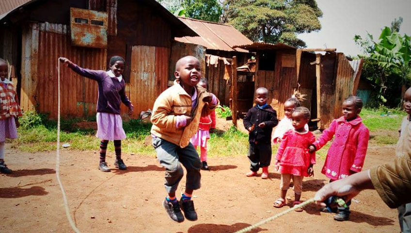 A group of children play outdoors in a rustic setting with a wooden shack in the background. One child in a brown jacket is mid-air, jumping rope while others watch and smile, their joyous expressions capturing the spirit of charity. The children are dressed in colorful clothing and stand on reddish dirt ground.