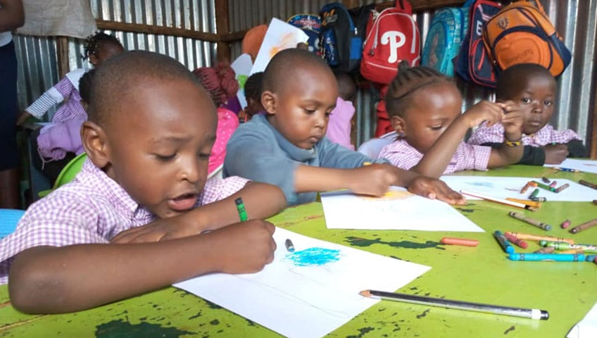 Four young children are seated at a green table, drawing and coloring on white sheets of paper with crayons and pencils. They are wearing checkered shirts, and colorful school bags hang behind them. The background features a metallic wall, highlighting the impact of the recent charity donation that made this creative space possible.