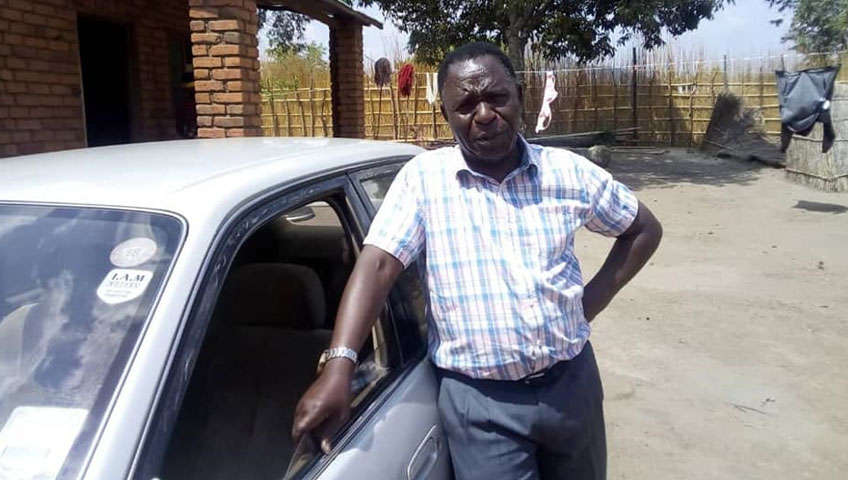 A man in a checkered shirt stands with his left hand resting on the roof of a silver car. He is positioned in front of a brick house, with various items hanging under a tree in the background. The scene suggests a rural setting where community efforts for water donation and charity are taking place.