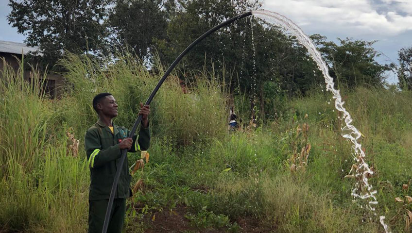 A man stands outdoors in a lush, green area, holding a thick black hose that arches upwards, releasing a strong stream of water. He wears a green uniform and appears to be irrigating the surrounding vegetation as part of a local charity initiative. Trees and a building are visible in the background.