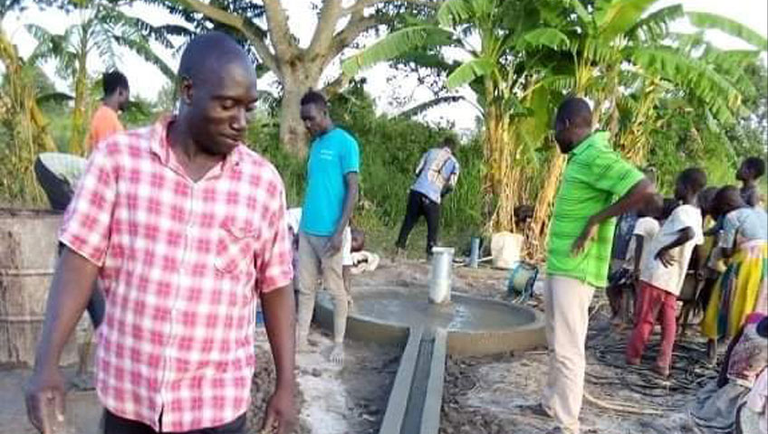 A group of people gather around a newly constructed water pump in a rural area, funded by a generous donation. One man in a pink plaid shirt stands in the foreground, while others, including children, are in the background near lush vegetation. The water pump has a spout with flowing water.