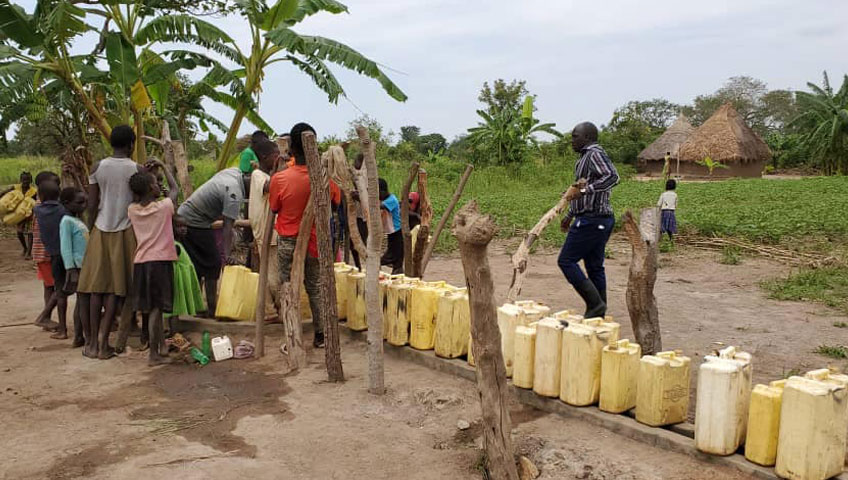 A group of people gathers around a communal water source in a rural setting, filling yellow jerry cans and containers supplied by a local charity. The background features lush green vegetation, banana trees, and a thatched-roof hut. Wooden posts line the area where the containers are being filled with clean water.