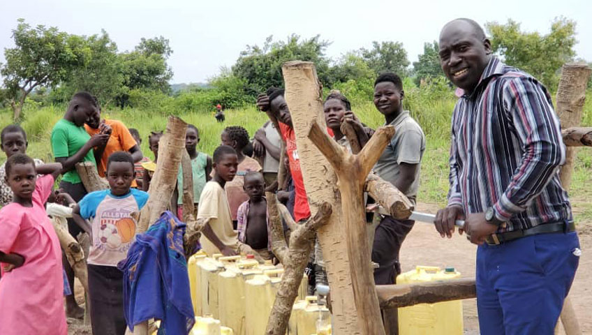 A group of children and adults gather around a water pump in a rural setting, filling yellow jerry cans. A man in a striped shirt and blue pants stands in the foreground, smiling at the camera. Trees and grassy fields are visible in the background, showcasing the impact of a recent charity donation.