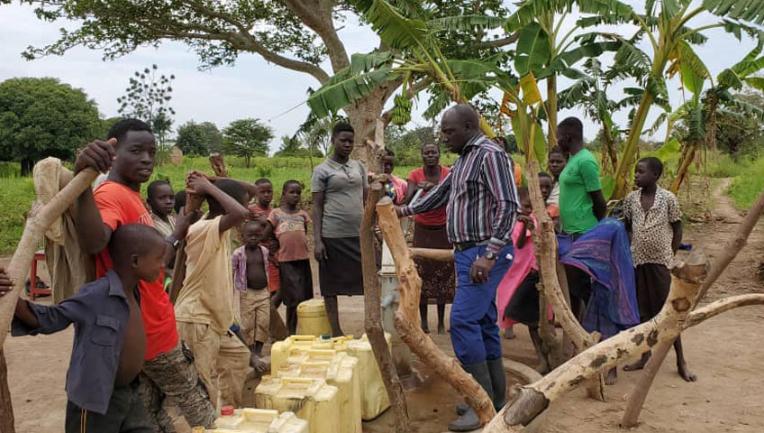 A group of people, including men, women, and children, gather around a makeshift water collection point in an outdoor setting with trees and vegetation in the background. Yellow water containers are scattered on the ground as a man in a striped shirt speaks to the group about the charity’s recent water donation.
