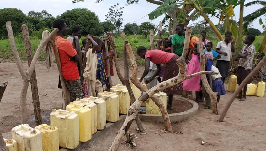 A group of people gathers around a water distribution point in a rural area. Several yellow jerry cans are lined up, with one person actively filling a container. Surrounding them are trees, and the background shows greenery and more people waiting—showcasing the impact of vital charity efforts.