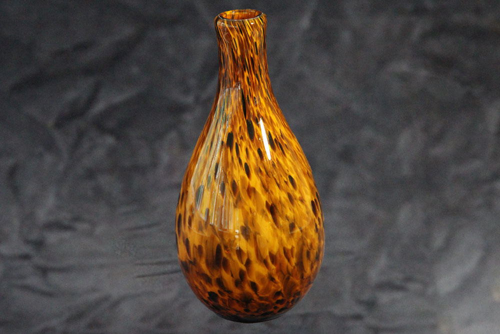 A teardrop-shaped glass vase with a narrow opening, featuring a striking pattern of amber and dark brown resembling a tiger's fur. The background is a blurred, dark surface that highlights the vase's intricate design, making it an elegant piece perfect for charity auctions or donation events.