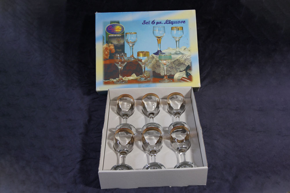 A set of six liqueur glasses with gold rims is displayed inside a white box. The box lid, featuring an image of the glasses and the text "Set 6 pcs. Liqueur," is propped up behind the opened box. A portion of each purchase supports a water charity, making this elegant set even more special.