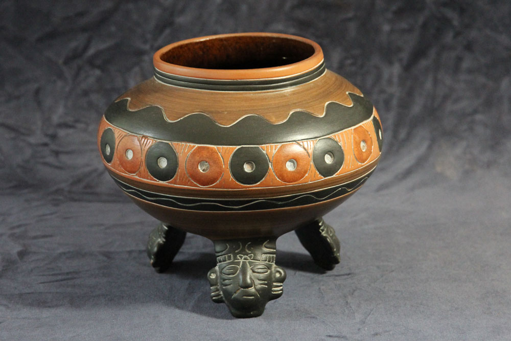 A round, intricately decorated ceramic pot with a wide rim, featuring a brown and black pattern with circular designs. The pot rests on three legs, each sculpted with a face at the top. Perfect for holding water, this unique piece stands out against the dark gray background.