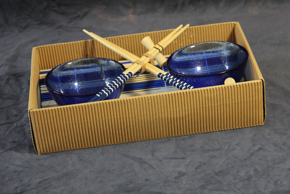 A rectangular gift box containing two blue bowls and two pairs of wooden chopsticks with blue and white striped handles, placed on a gray surface, makes a perfect item to donate to charity.