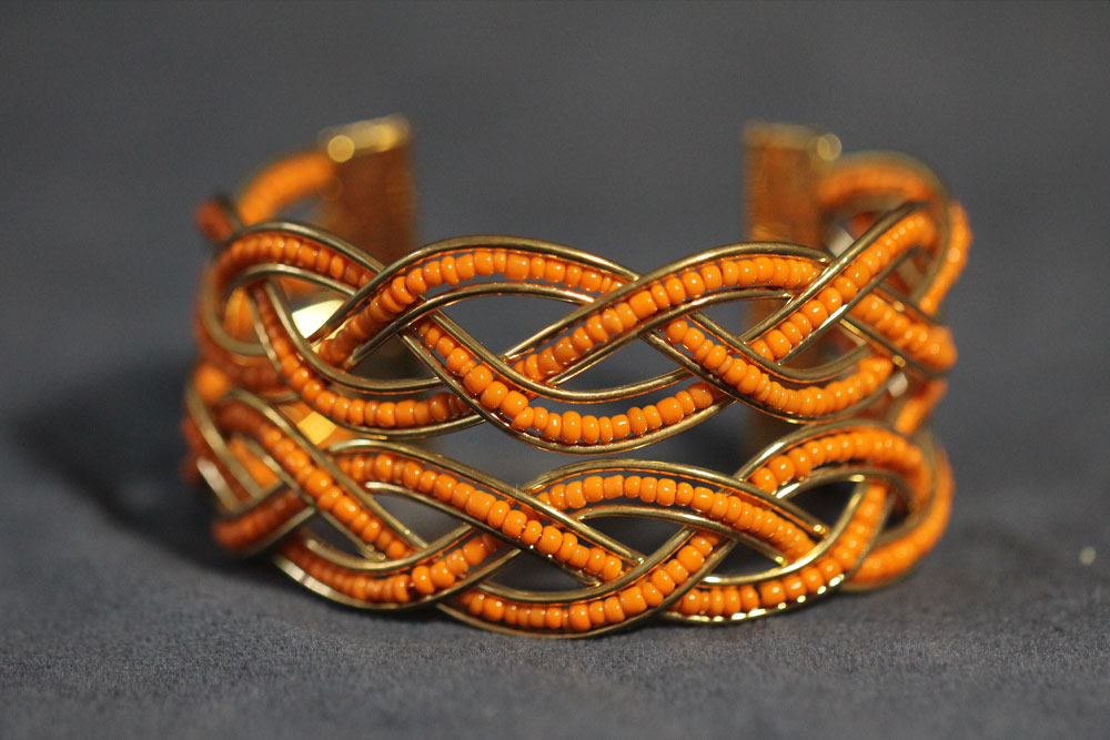 A close-up of a gold cuff bracelet intricately designed with multiple intertwining loops of orange beads, symbolizing charity. The bracelet is set against a plain dark background.