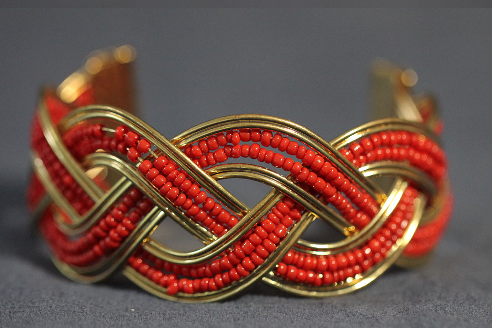 A gold bracelet intricately woven with strands of small red beads, creating a bold and elegant intertwined pattern reminiscent of flowing water. The design is detailed and symmetrical, giving the bracelet a sophisticated and artisanal look. The background is a plain, dark surface.